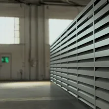 Interior of an industrial warehouse with large metal louvered panels on the right and bright windows in the background.