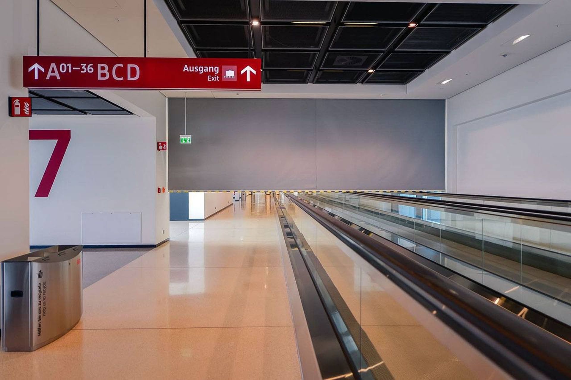 Empty airport hallway with a moving walkway, red exit signs, and a large number seven on the wall. A Smokemaster SM5 is installed above everything