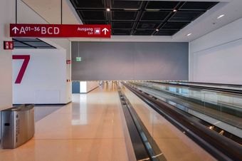 Empty airport terminal with a smoke curtain. Features include a moving walkway, red signs indicating exits and gates A01-36, a large "7" on the wall, and a trash bin in the corner.