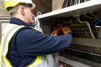 An electrician in a safety vest and helmet uses a multimeter to check wires inside an electrical panel.