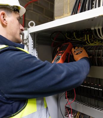 An electrician in a safety vest and helmet uses a multimeter to check wires inside an electrical panel.
