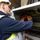 An electrician in a safety vest and helmet uses a multimeter to check wires inside an electrical panel.