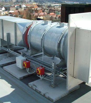 Industrial ventilation unit on a rooftop with a cityscape in the background. The unit consists of ducts, a fan, and control boxes.