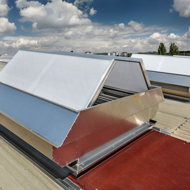 Rooftop view of metal ventilation units against a cloudy sky, with a mix of beige and red roofing panels.