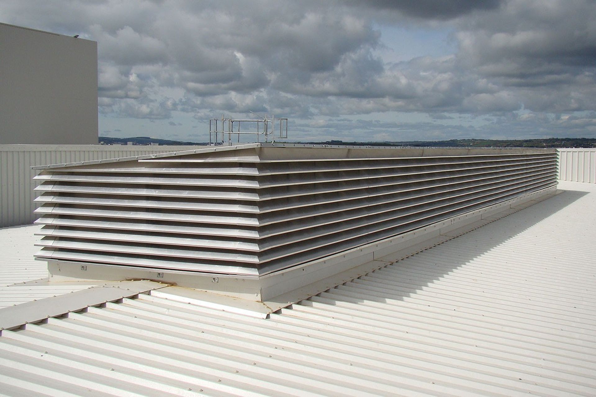 Large rooftop air vent with horizontal metal slats on a white corrugated surface under a partly cloudy sky.