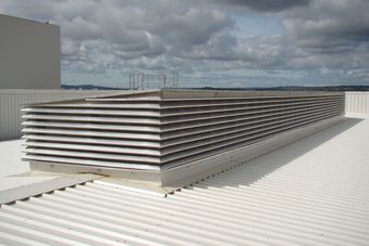 Large rooftop air vent with horizontal metal slats on a white corrugated surface under a partly cloudy sky.