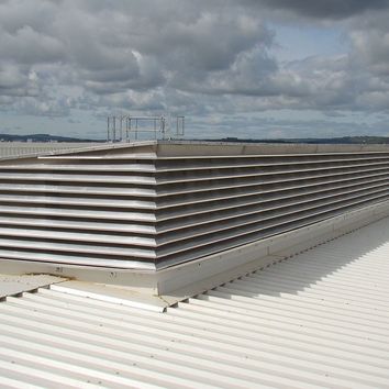 Large rooftop air vent with horizontal metal slats on a white corrugated surface under a partly cloudy sky.
