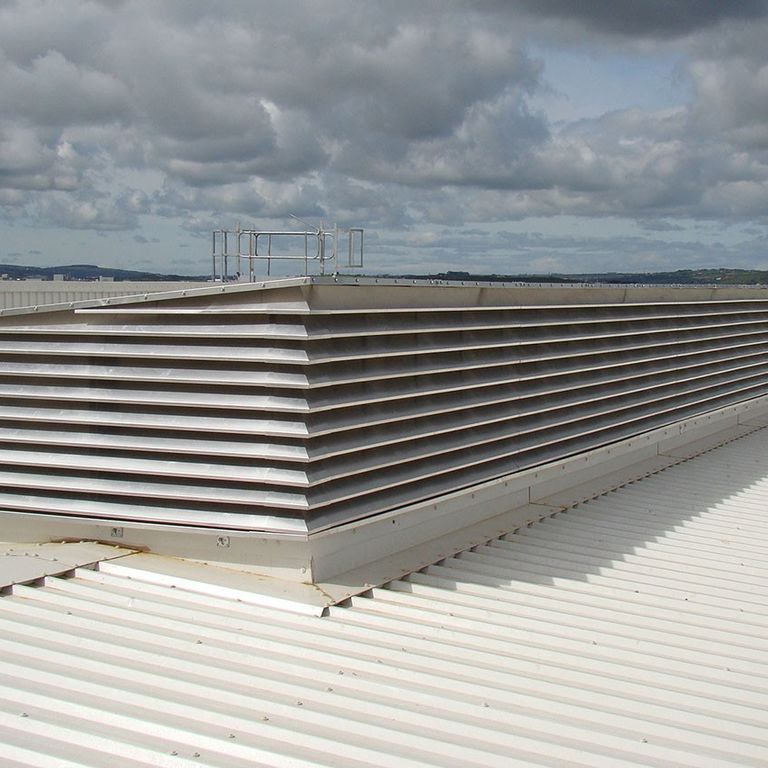 Large rooftop air vent with horizontal metal slats on a white corrugated surface under a partly cloudy sky.