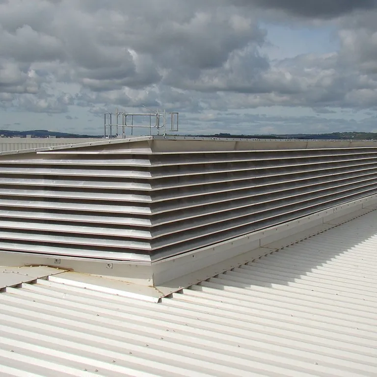 Large rooftop air vent with horizontal metal slats on a white corrugated surface under a partly cloudy sky.