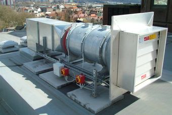 Rooftop industrial ventilation system with metal ducts and control boxes, overlooking a cityscape with buildings and trees in the background.