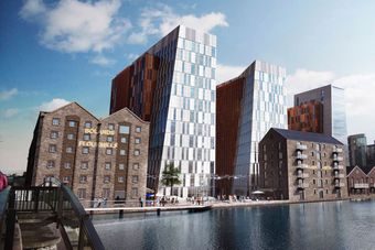 Modern glass buildings and historic brick warehouses line a waterfront, with people walking along the promenade under a blue sky.