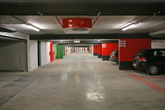 Empty underground parking garage with red and white walls, a few cars parked on the right, and green and red emergency exit doors.