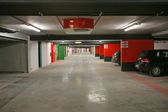 Empty underground parking garage with red and white walls, a few cars parked on the right, and green and red emergency exit doors.