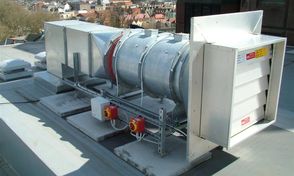 Industrial rooftop ventilation unit with a cylindrical metal duct and square outlets, overlooking city buildings in the background.