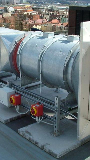 Industrial rooftop ventilation unit with a cylindrical metal duct and square outlets, overlooking city buildings in the background.