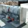 Industrial rooftop ventilation unit with a cylindrical metal duct and square outlets, overlooking city buildings in the background.