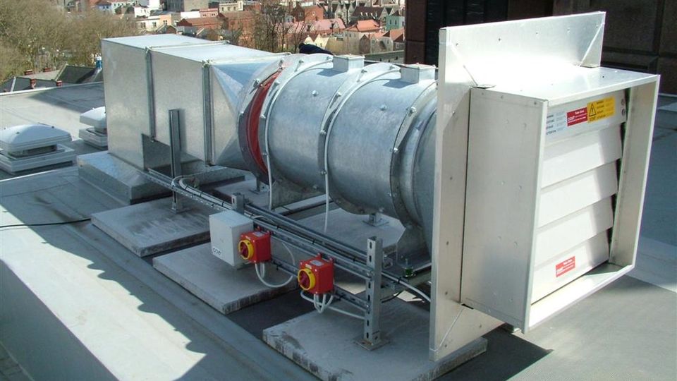 Industrial rooftop ventilation unit with a cylindrical metal duct and square outlets, overlooking city buildings in the background.