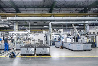 Interior of an industrial factory with machinery, metal ducts, and large crates labeled BMW. Two workers are seen moving amidst the complex setup.