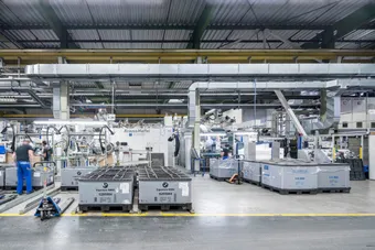 Interior of an industrial factory with machinery, metal ducts, and large crates labeled BMW. Two workers are seen moving amidst the complex setup.
