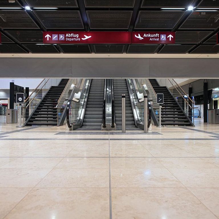 Sleek airport interior with escalators and stairs under signs for departures and arrivals in multiple languages. A Smokemaster SM5 is installed above the escalator.