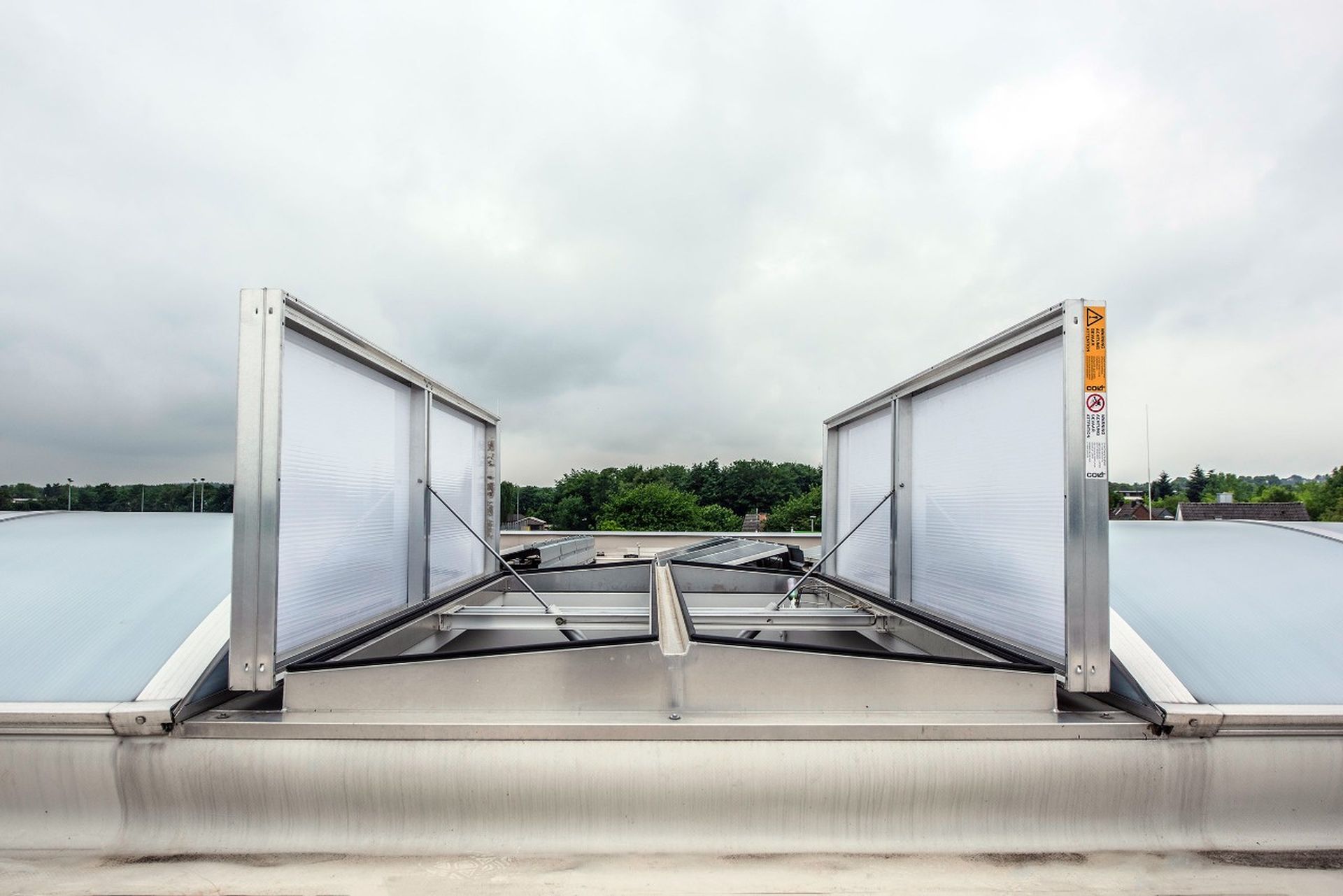 Skylight with open vents on a rooftop, surrounded by greenery and a cloudy sky in the background.