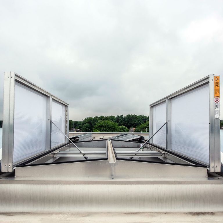 Skylight with open vents on a rooftop, surrounded by greenery and a cloudy sky in the background.