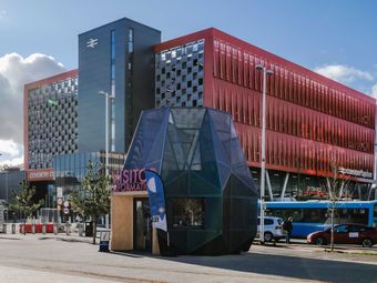 Modern urban landscape with Coventry Station's red building, a unique geometric visitor information booth, and street activity under a blue sky.