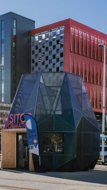 Modern urban landscape with Coventry Station's red building, a unique geometric visitor information booth, and street activity under a blue sky.