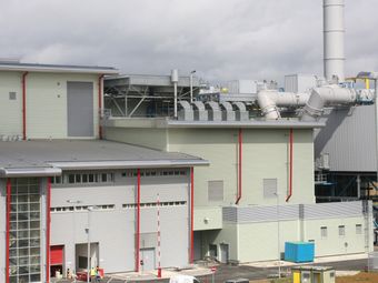 Exterior view of an industrial building with red accents, large ventilation pipes, and a tall chimney under a cloudy sky.