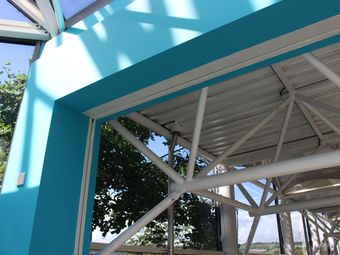 Interior view of a modern building with a turquoise beam, metal trusses, and large windows revealing trees and blue sky outside.
