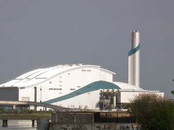 A large industrial building with a curved roof and a tall chimney, featuring a blue wave design, situated near a body of water.