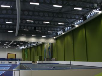 Indoor sports hall with blue tennis courts, green walls, and a climbing wall, illuminated by ceiling lights and natural light from high windows.