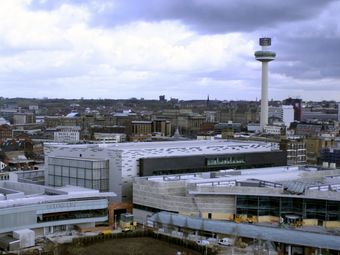 Aerial view of Liverpool with modern and historical buildings, featuring a tall radio tower and overcast sky in the background.