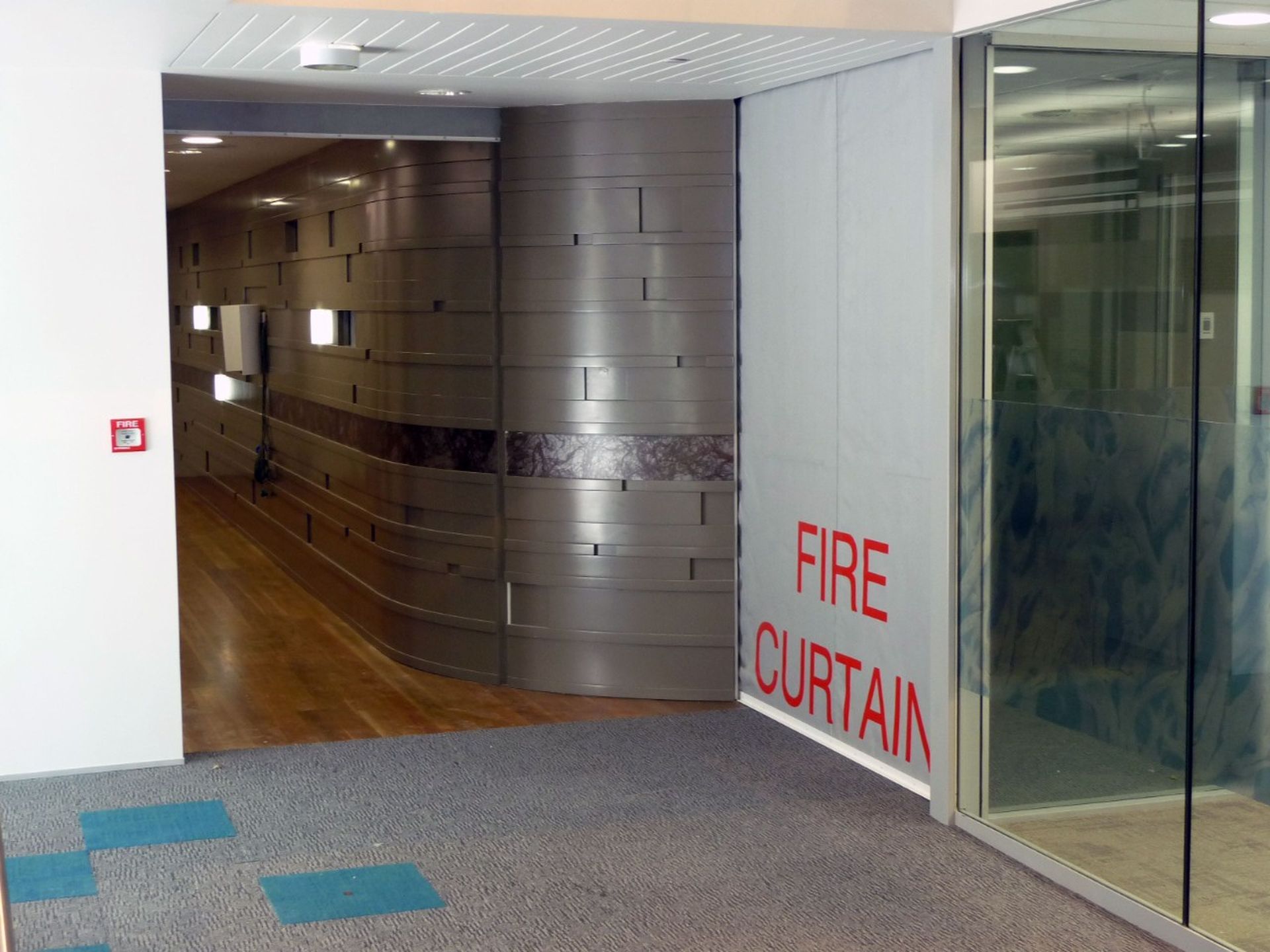 Corridor with metallic wall and wooden floor, beside a glass section. A Fire Curtain FM1 is partially visible with a nearby fire alarm.
