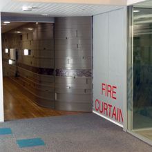Corridor with metallic wall and wooden floor, beside a glass section. A Fire Curtain FM1 is partially visible with a nearby fire alarm.