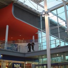 Modern indoor shopping mall with large glass windows, a red ceiling, and people walking on an upper-level walkway.