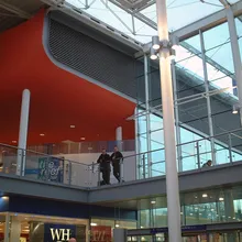 Modern indoor shopping mall with large glass windows, a red ceiling, and people walking on an upper-level walkway.