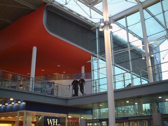 Modern indoor shopping mall with large glass windows, a red ceiling, and people walking on an upper-level walkway.
