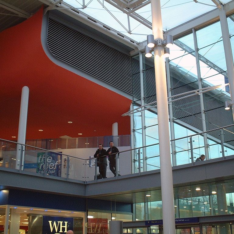 Modern indoor shopping mall with large glass windows, a red ceiling, and people walking on an upper-level walkway.