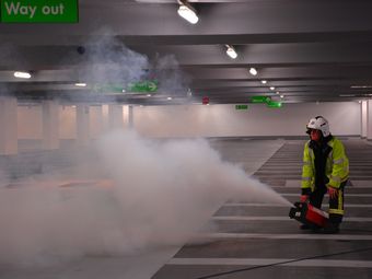A firefighter in uniform operates a smoke machine in a parking garage, with "Way Out" signs visible and smoke filling the area.