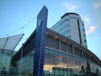 Manchester Piccadilly Station, a modern glass and steel building with a tall blue sign and curved roof, reflecting a sunset sky in its glass facade.