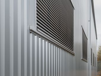 Exterior wall of a metal building with vertical grooves and large louvered vents, viewed from a side angle.