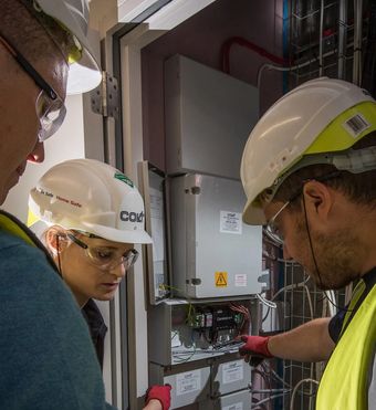 Three technicians wearing hard hats and safety gear inspect electrical equipment in an industrial setting.