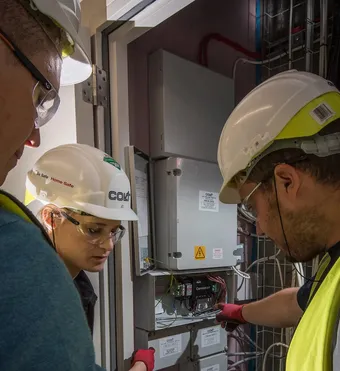 Three technicians wearing hard hats and safety gear inspect electrical equipment in an industrial setting.