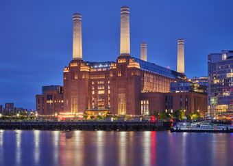 Illuminated historic brick building with four tall chimneys by a river at night, reflecting lights on the water.