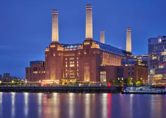 Illuminated historic brick building with four tall chimneys by a river at night, reflecting lights on the water.