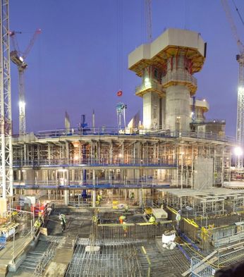 Construction site at dusk, bustling with workers and scaffolding. Tall cranes surround the multi-level structure, illuminated by bright lights.