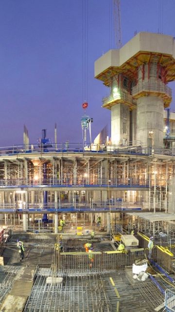 Construction site at dusk, bustling with workers and scaffolding. Tall cranes surround the multi-level structure, illuminated by bright lights.