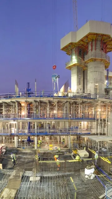 Construction site at dusk, bustling with workers and scaffolding. Tall cranes surround the multi-level structure, illuminated by bright lights.