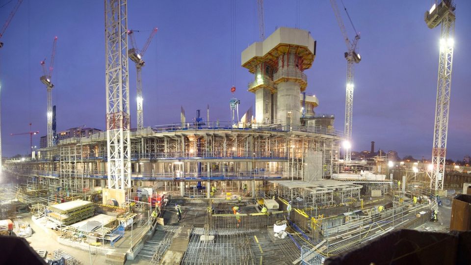 Construction site at dusk, bustling with workers and scaffolding. Tall cranes surround the multi-level structure, illuminated by bright lights.